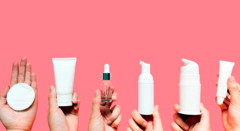Multiple hands holding various white skincare tubes and bottles against a bright pink background.