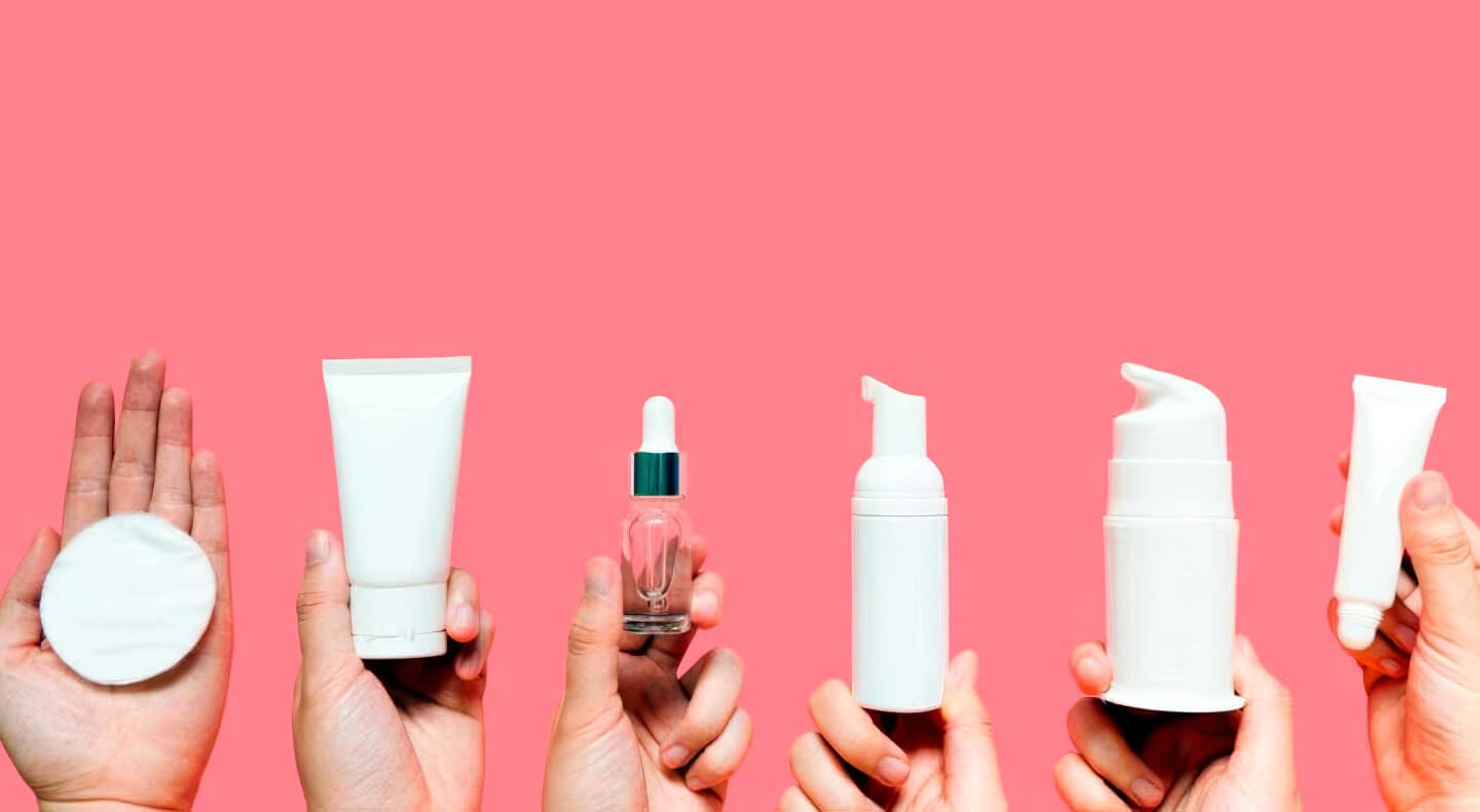 Multiple hands holding various white skincare tubes and bottles against a bright pink background.