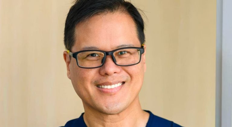 A smiling doctor with glasses wearing blue clinical scrubs.