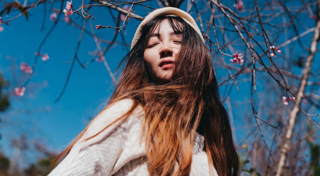 A woman with her eyes closed enjoying the sun under pink cherry blossom trees.