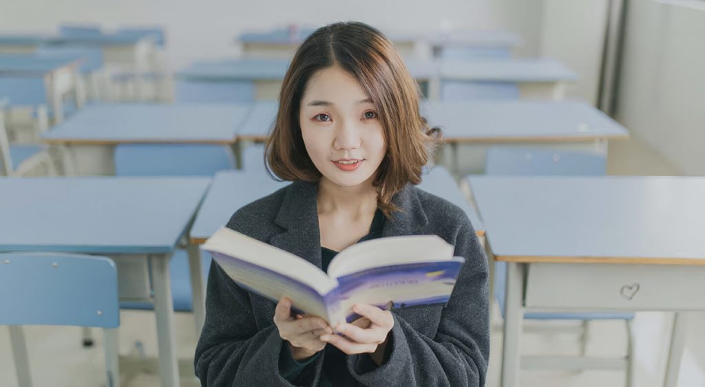 A woman sitting in a classroom or library setting holding an open book and looking at the camera.