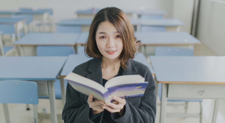 A woman sitting in a classroom or library setting holding an open book and looking at the camera.